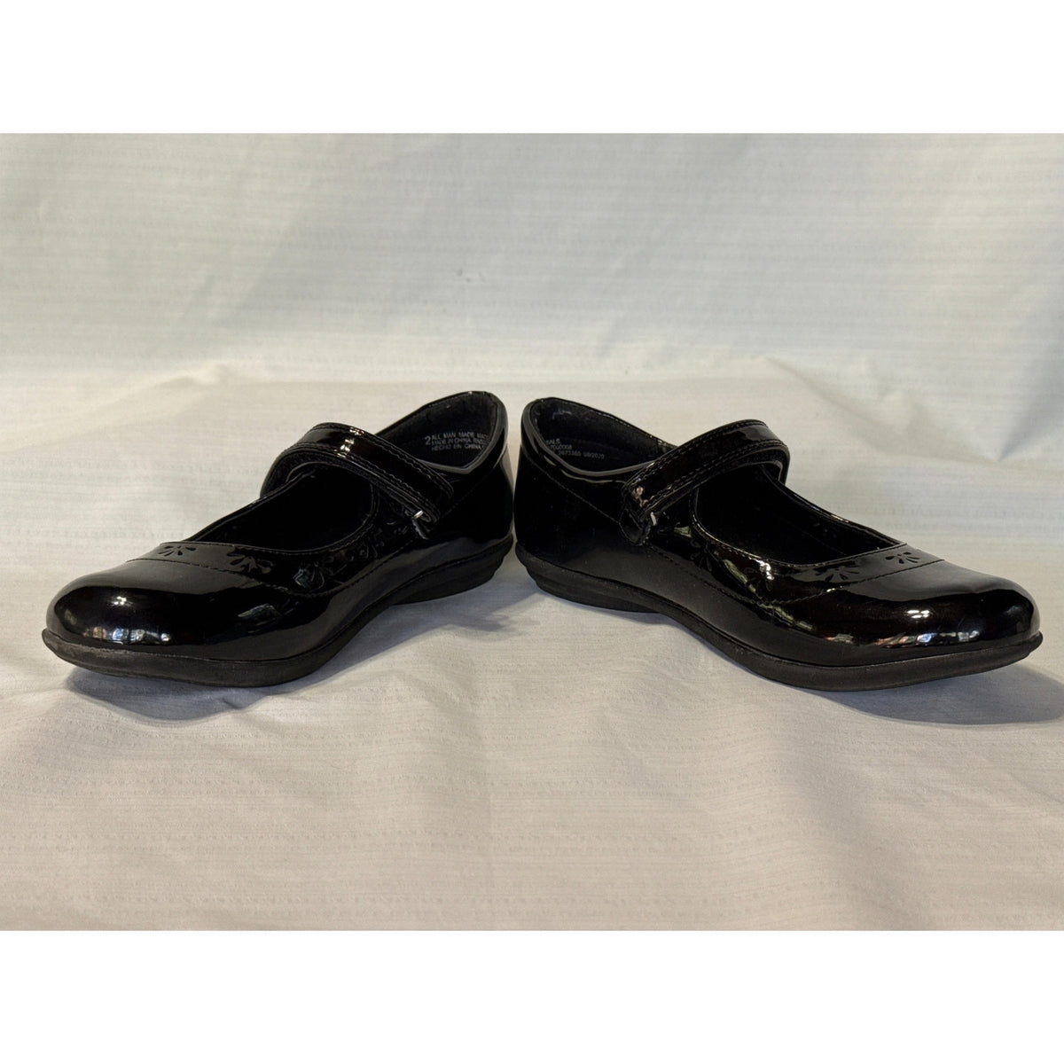 Angled front view of The Children’s Place black Mary Jane shoes showing shiny uppers, straps, and floral cutout trim.