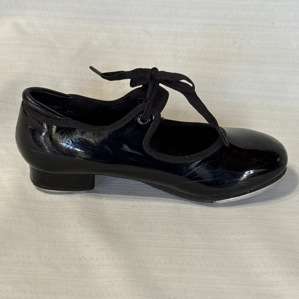 Side view of a black Balera tie tap shoe showing scuffs near the heel and a low block heel.
