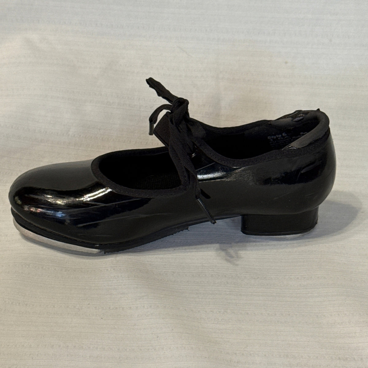 Side profile of a glossy black Balera tap shoe with a round toe, bow tie, and low heel.
