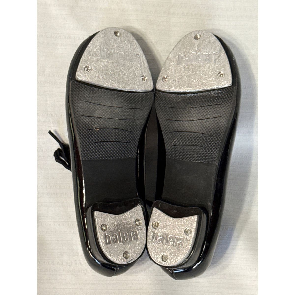 Bottom view of black Balera tap shoes showing textured rubber soles and worn metal toe and heel taps.
