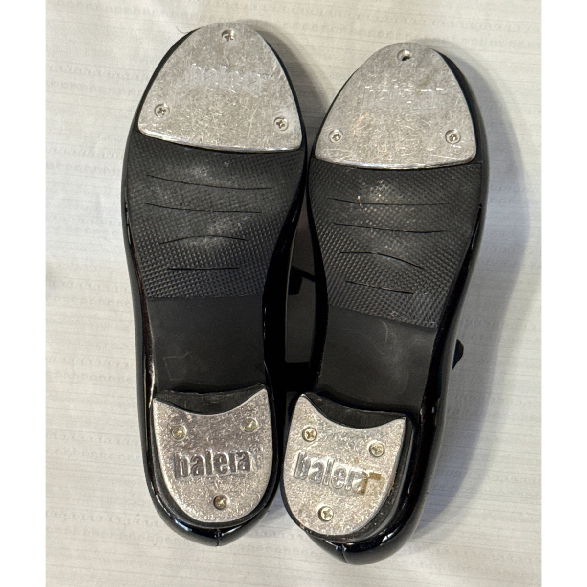 Soles of Balera black tap shoes showing worn metal toe and heel taps, with one screw missing from the left toe tap.
