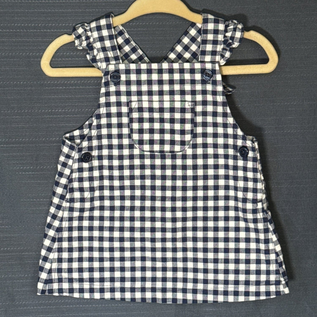 Front view of Carter's Child of Mine baby girls' gingham pinafore dress in navy blue and white, featuring ruffled shoulders, front pocket, and decorative buttons.