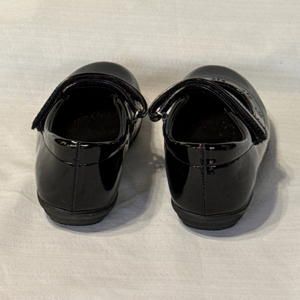 Back view of girls’ black Mary Jane shoes showing heel seams, glossy finish, and low heels.
