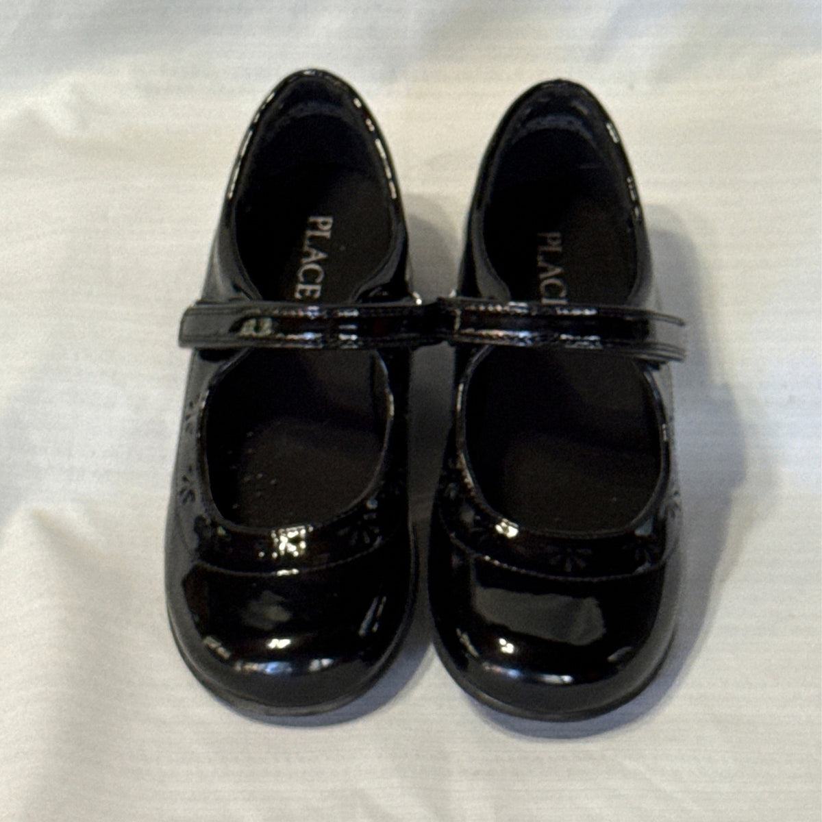 Front view of The Children’s Place black Mary Jane shoes showing rounded toes, instep straps, and flower-shaped cutouts.