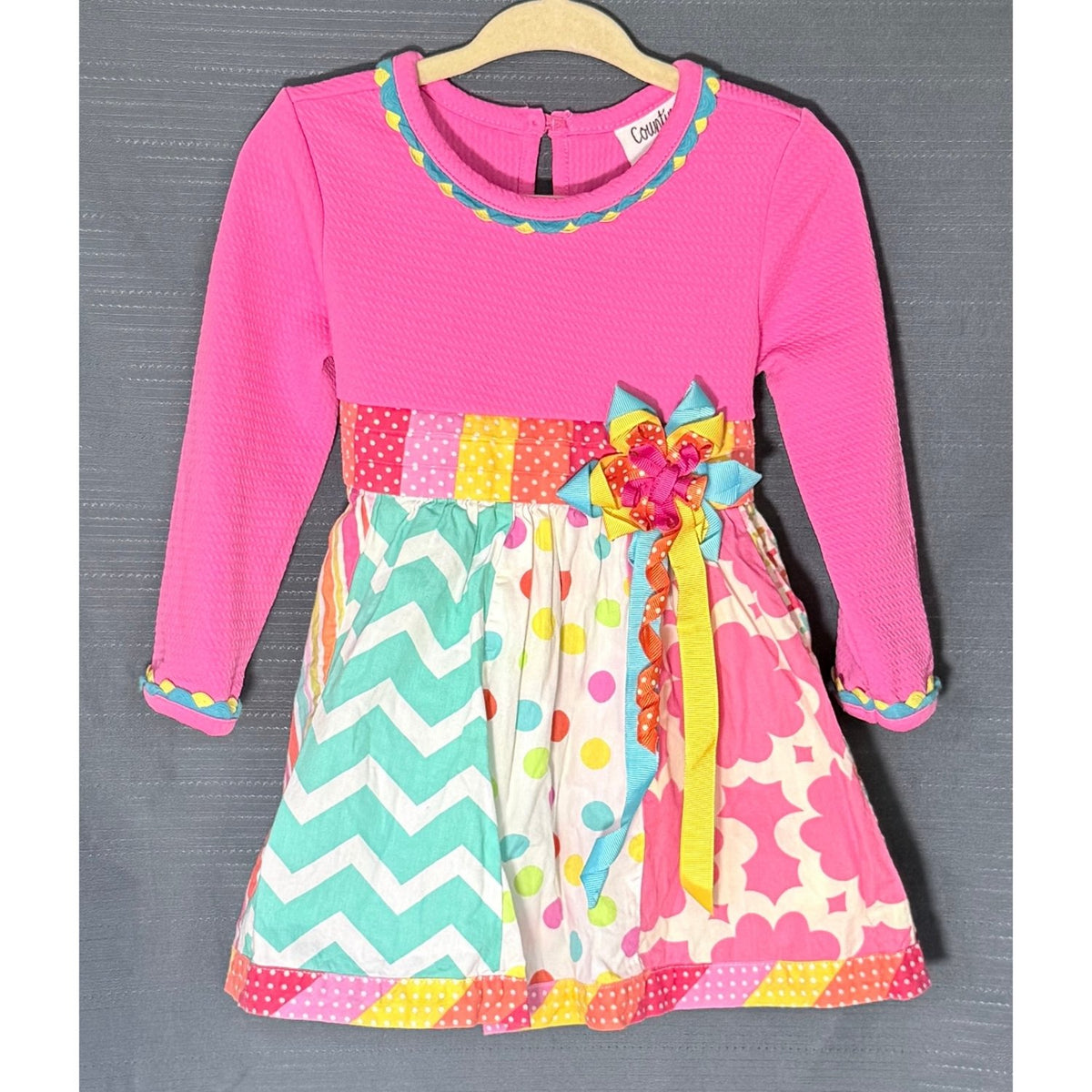 Front view of Counting Daisies 18-month dress with pink textured top, colorful patchwork-style skirt, and decorative ribbon bow.