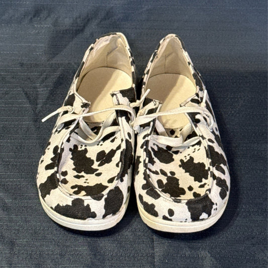 Front view of black-and-white cow-print shoes showing rounded toes, moc-toe stitching, and white laces.