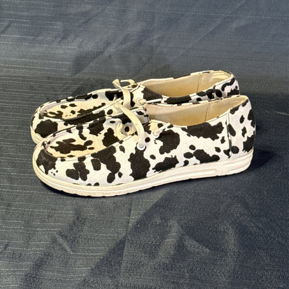 Left-side view of cow-print canvas slip-on shoes showing the low-cut shape and light-colored soles.
