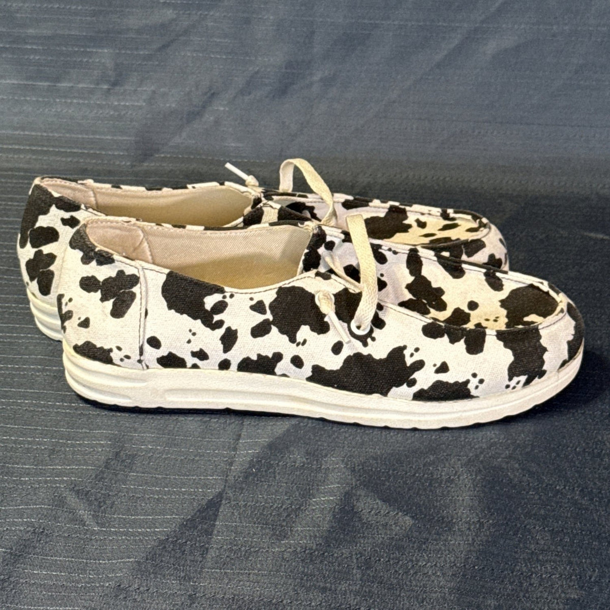 Right-side view of black-and-white cow-print slip-on shoes with white laces and stitched toe panels.
