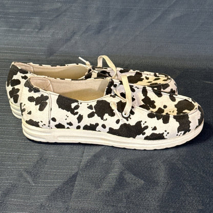 Right-side view of black-and-white cow-print slip-on shoes with white laces and stitched toe panels.
