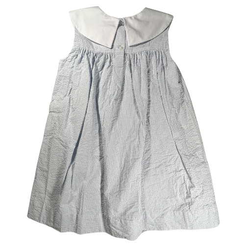 The image shows the back view of a sleeveless, knee-length girl's dress. The dress is made of light blue fabric with a seersucker texture and a subtle windowpane pattern. The back features a white yoke with a pointed collar, secured by two buttons at the neckline. The fabric is gathered just below the yoke, creating a gently flared silhouette that flows down to the hem. The overall design is simple yet elegant, with a light and airy feel. Shop at ourfamiliesattic.com