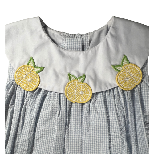 The image is a close-up of the neckline of a girl's dress, showcasing the white yoke and collar. The yoke is adorned with three lemon appliqués, each featuring a bright yellow lemon slice with green leaves. The lemons are evenly spaced across the front of the yoke, adding a playful and cheerful touch to the dress. The background fabric is light blue with a seersucker texture and a subtle windowpane pattern. The collar has a gentle curve and complements the overall design, highlighting the fresh and summery.