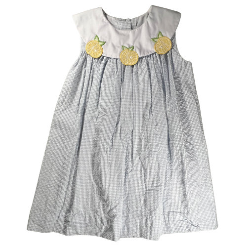 The image shows a sleeveless, knee-length dress designed for a young girl. The dress features a light blue and white seersucker pattern, characterized windowpane pattern. The bodice has a white yoke with three yellow lemon appliqués stitched across the front, each with a small green leaf attached. The dress has a round neckline, and the fabric flows gently from the yoke, giving it a slightly flared silhouette. Our Families Attic.