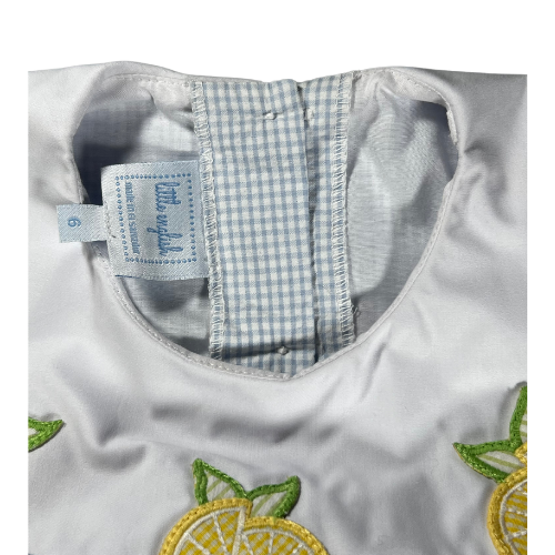 The image is a close-up of the label of a girl's dress, showcasing the white yoke and collar. The yoke is adorned with three lemon appliqués, each featuring a bright yellow lemon slice with green leaves. The lemons are evenly spaced across the front of the yoke, adding a playful and cheerful touch to the dress. The background fabric is light blue with a seersucker texture and a subtle windowpane pattern. The collar has a gentle curve and complements the overall design, highlighting the fresh and summery.