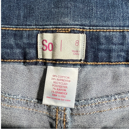 Tag of Gently used blue jean overalls for big girls size 8, by So, featuring snap sides, 5 pockets, rolled-up legs, and belt loops. Made from 68% cotton, 24% recycled polyester, 7% rayon, and 1% spandex. Our Families Attic