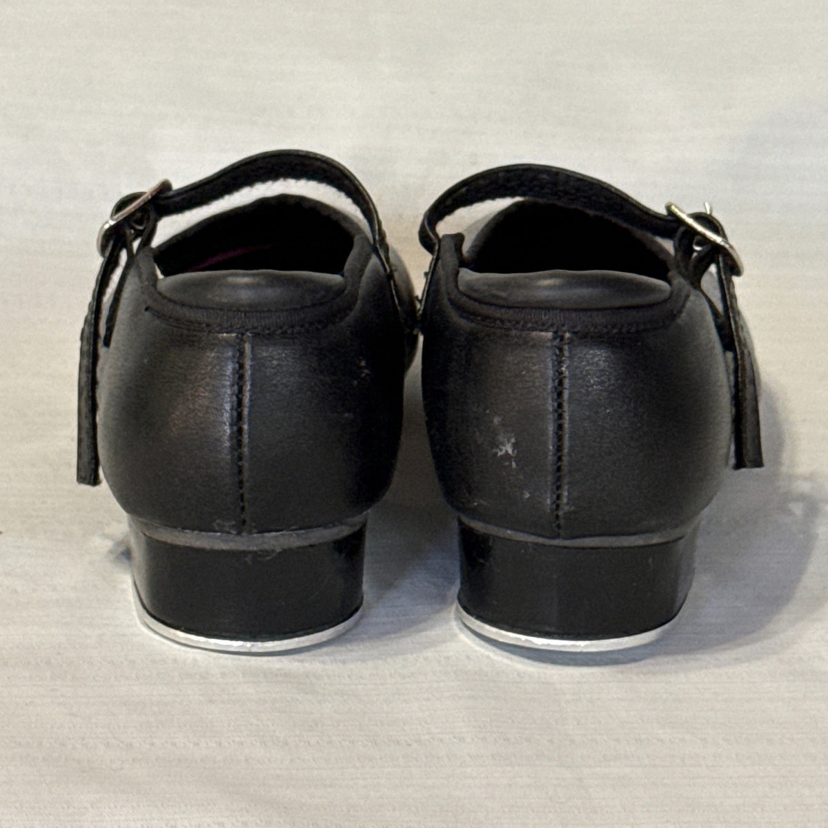 Back view of Revolution black tap shoes showing heel seams, light scuffs, buckle straps, and low heels.
