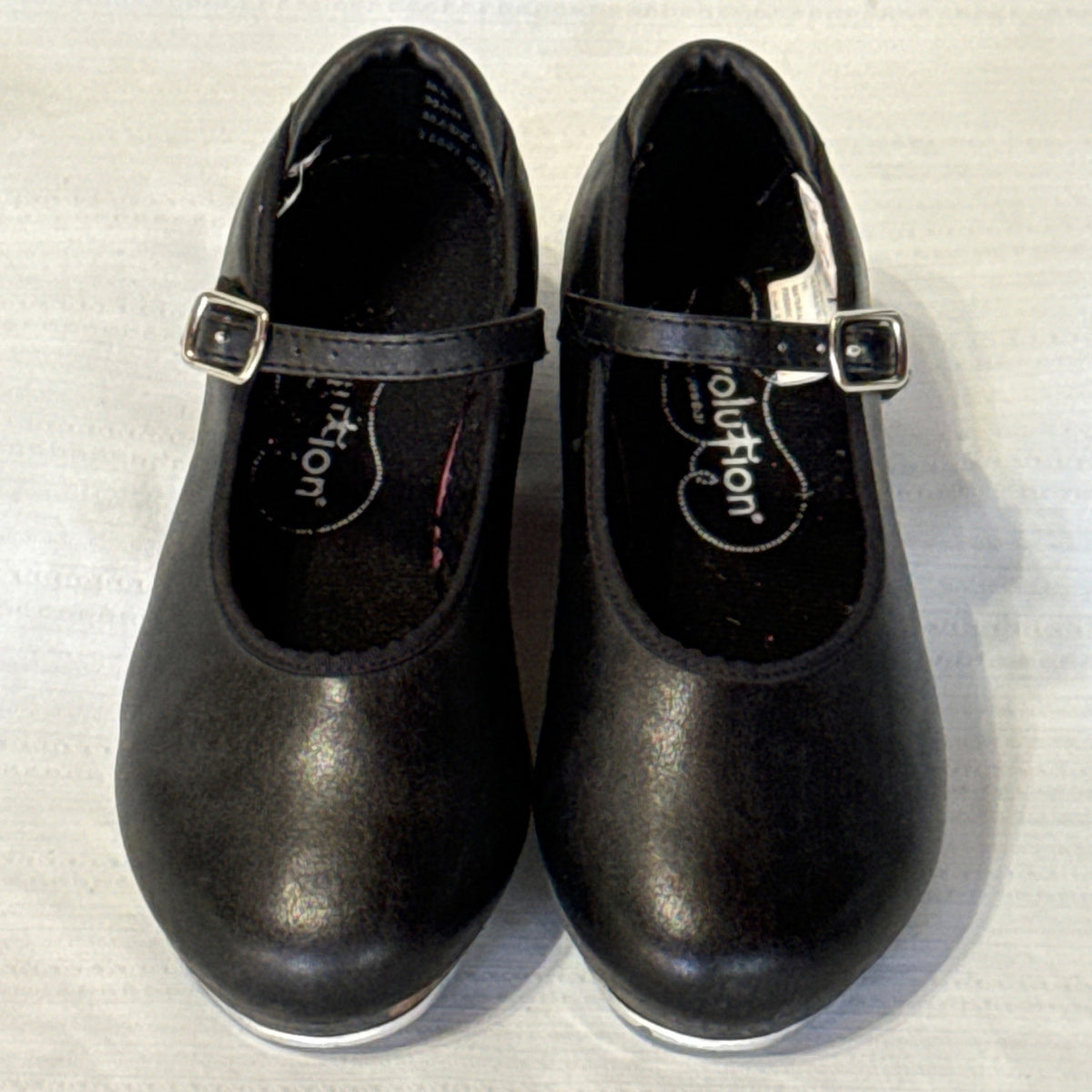 Front view of Revolution black buckle tap shoes showing rounded toes and lightly textured uppers.