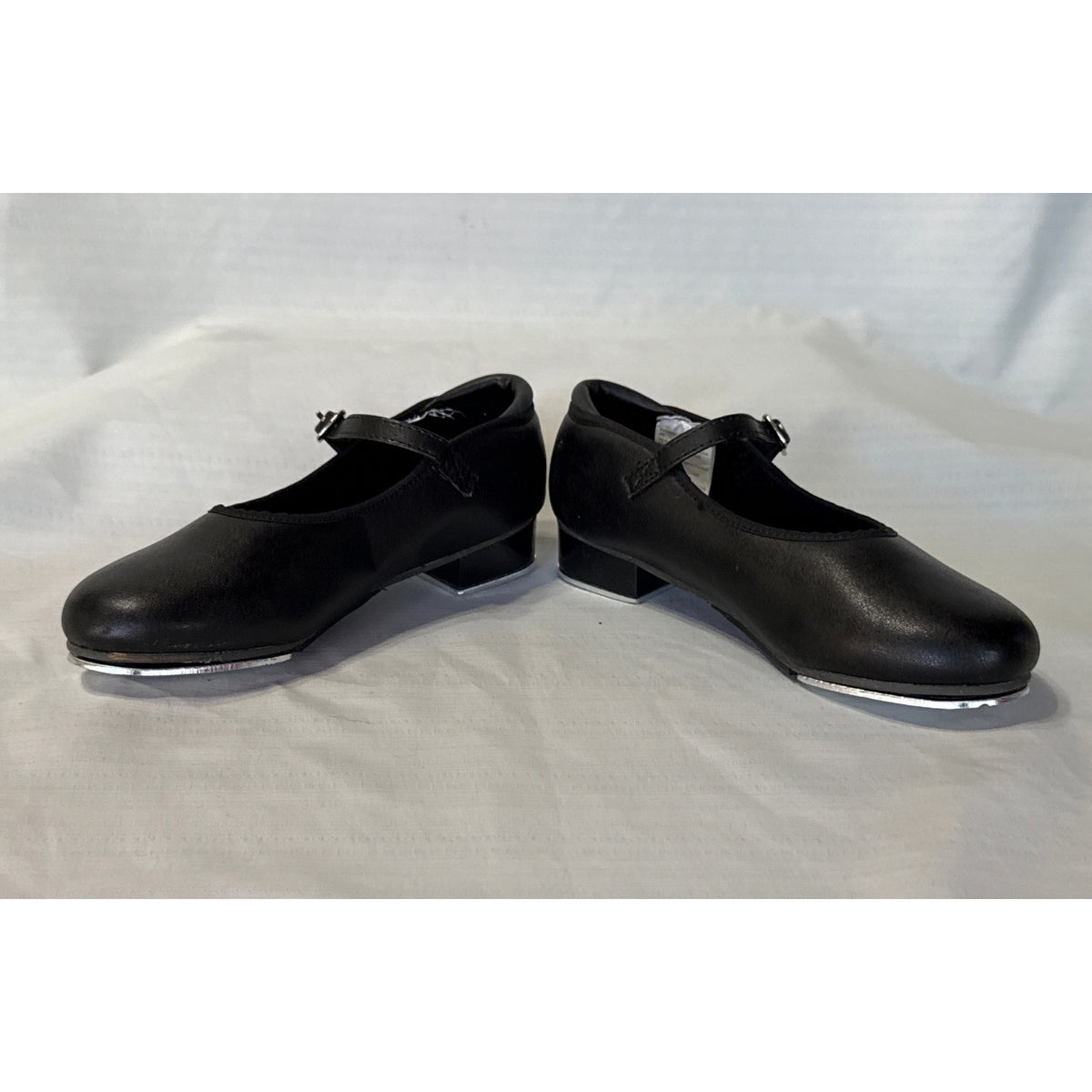 Angled side view of Revolution black Mary Jane tap shoes showing round toes, buckle straps, and low block heels.
