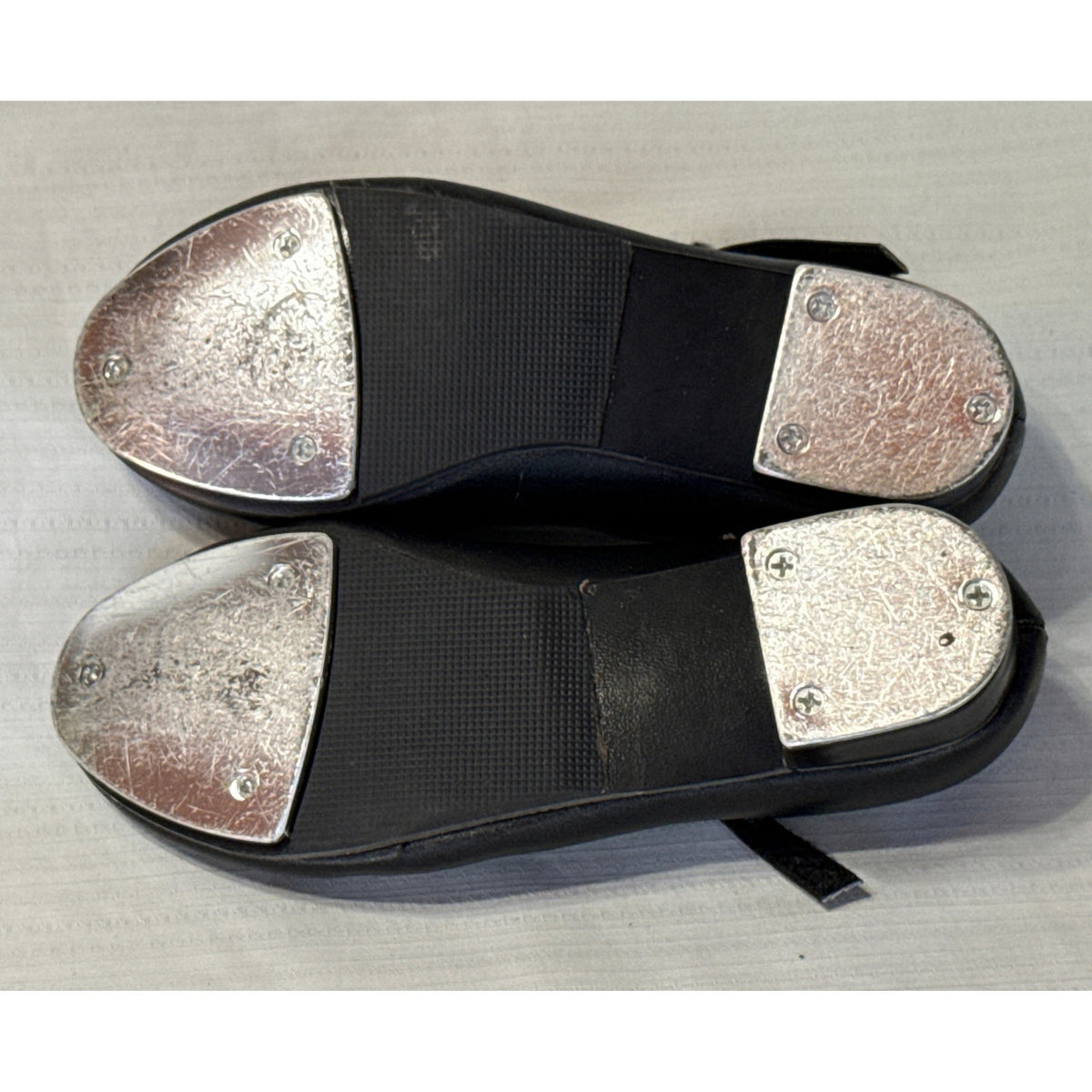 Bottom view of Revolution black tap shoes showing textured soles and scratched metal toe and heel taps.
