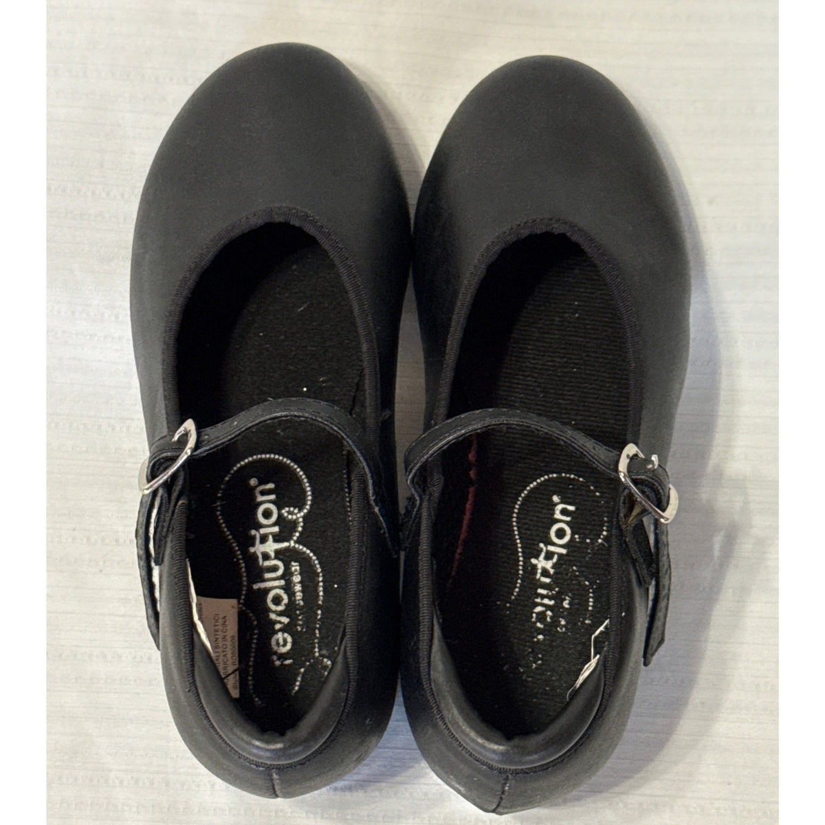 Top view of Revolution black tap shoes showing adjustable buckle straps, branded insoles, and an inside material label.
