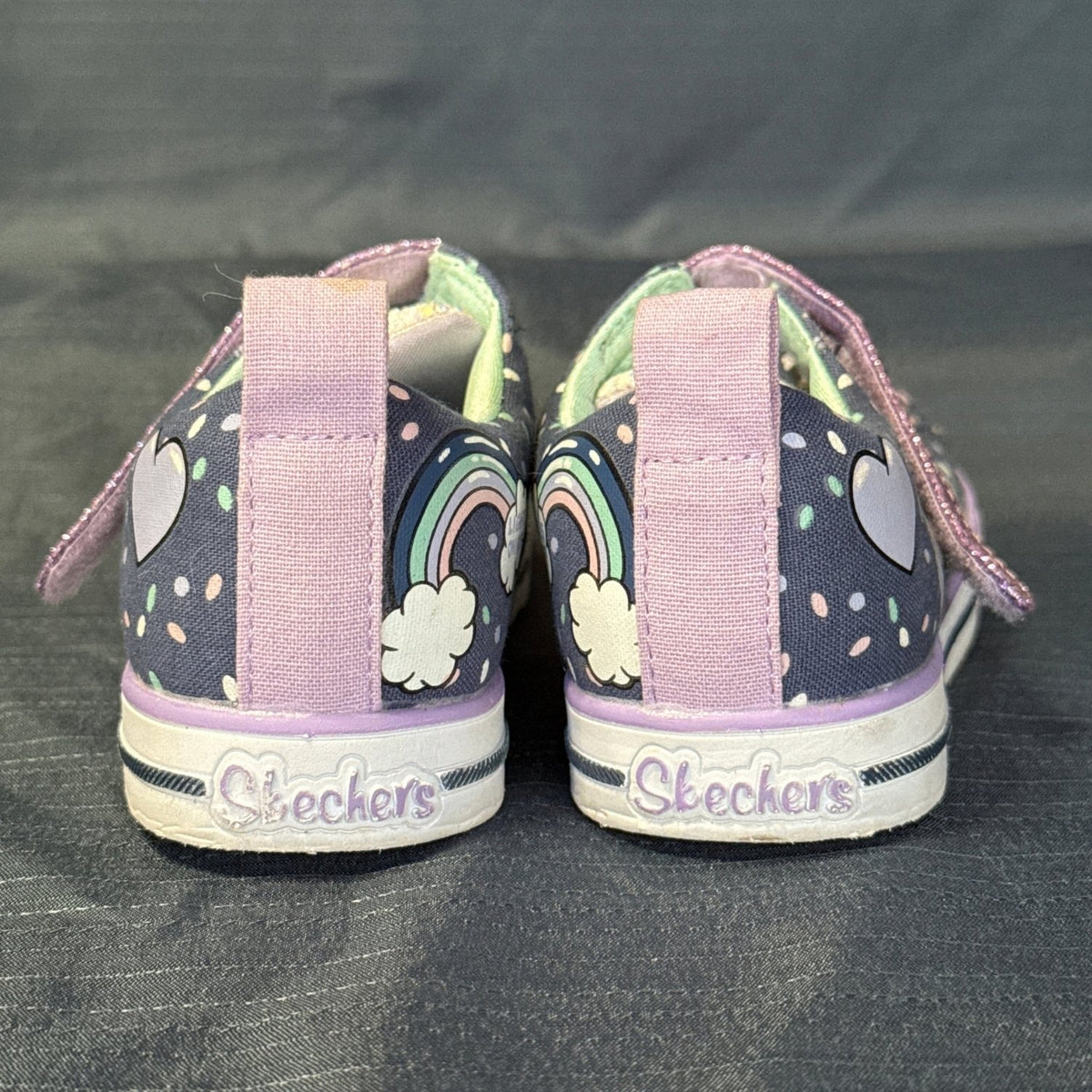 Back view of Skechers Twinkle Toes sneakers with purple pull tabs, rainbow graphics, and Skechers heel branding.
