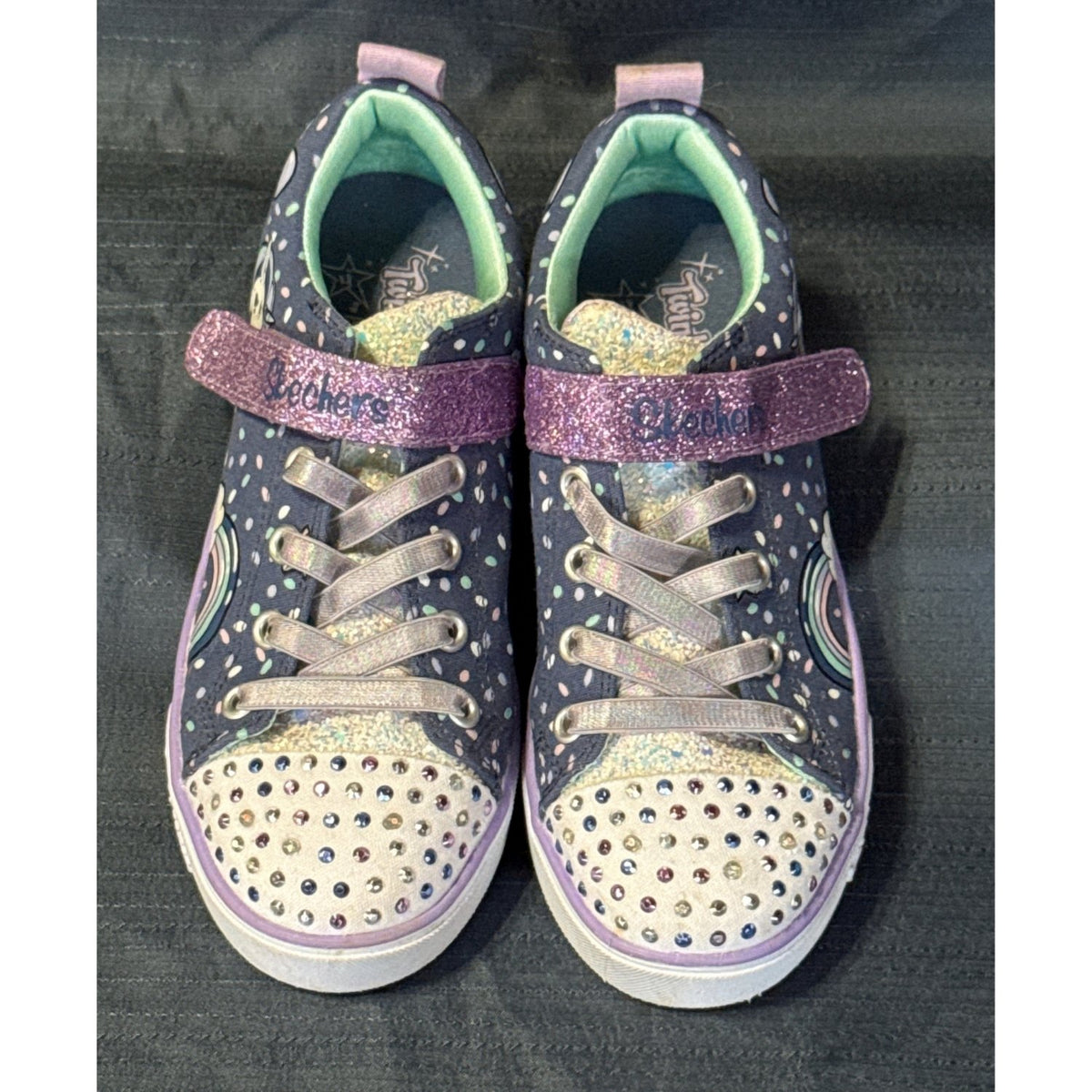 Front and top view of Skechers Twinkle Toes sneakers showing purple glitter straps, sequin tongues, stretchy laces, and multicolored toe gems.