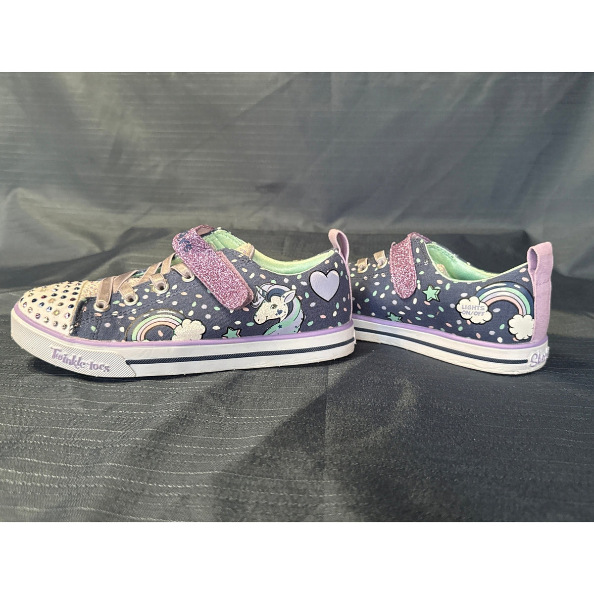 Side view of navy Skechers Twinkle Toes sneakers with unicorn and rainbow designs, purple glitter straps, and gem-covered toes.
