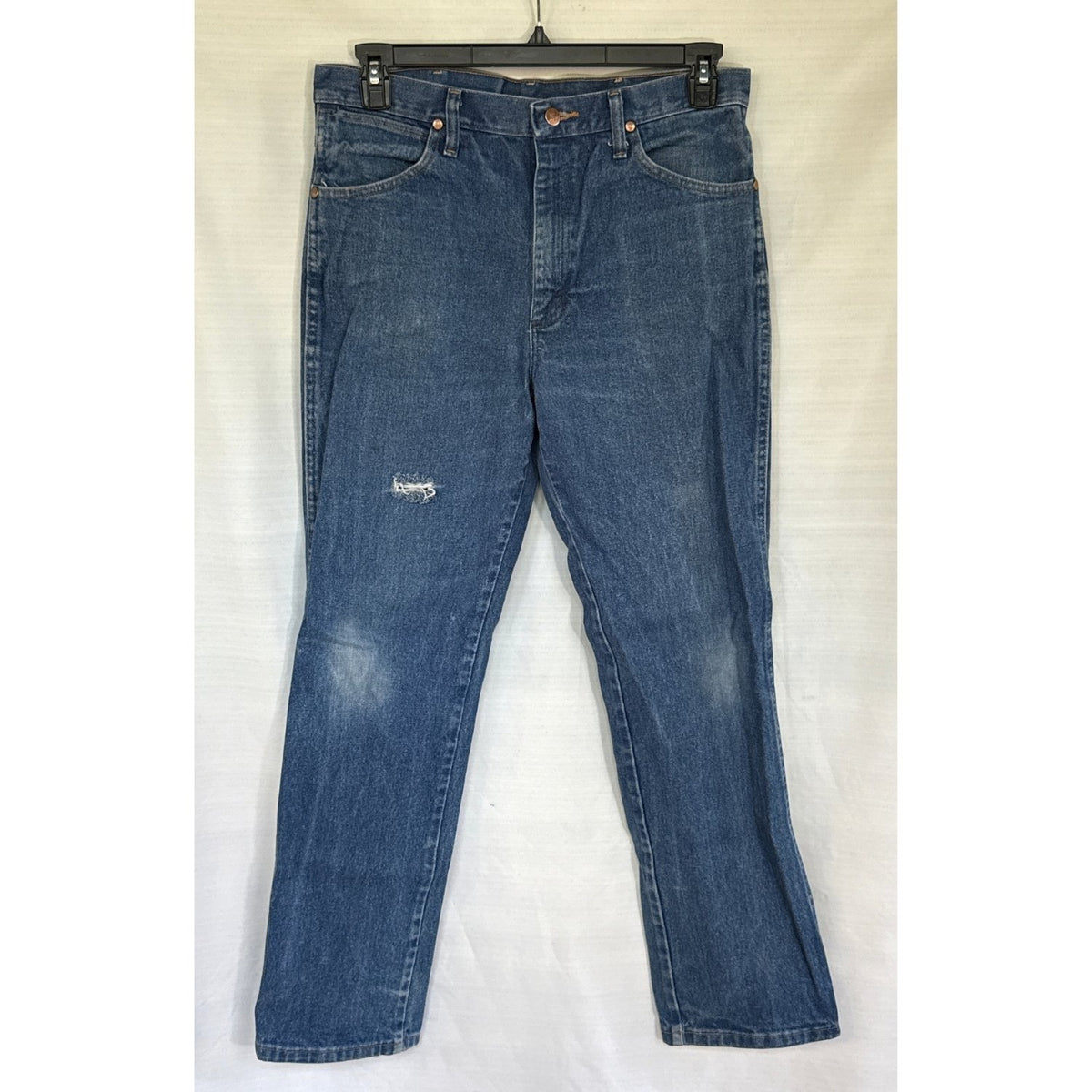 Front view of blue Wrangler men’s jeans with fading and a small rip on the right leg above the knee